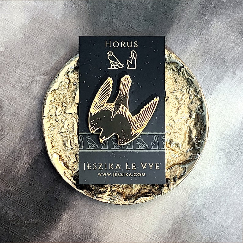 May include: A gold-colored enamel pin featuring a stylized depiction of the Egyptian god Horus, with the text "HORUS" and "JESZIKA LE VYE" and the website address "www.jeszika.com" on a black background.