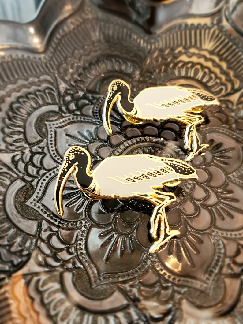 May include: Two gold-colored enamel pins of a white ibis bird with black accents. The pins are on a dark gray background with a decorative pattern.