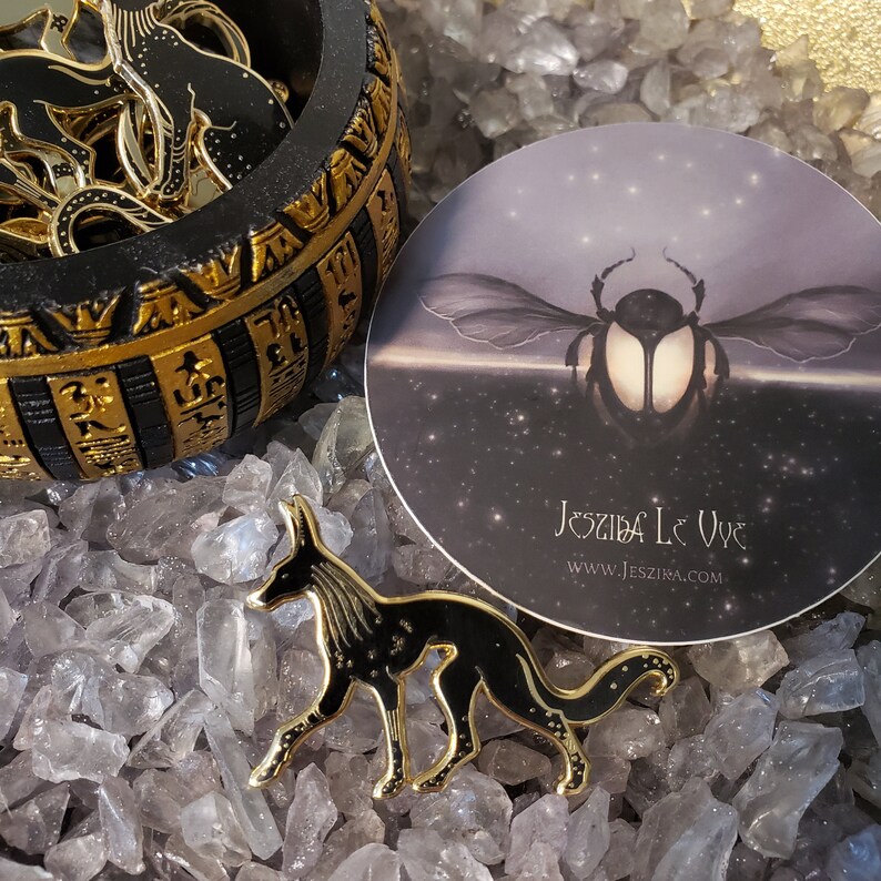 May include: Gold-toned enamel pin featuring a black stylized jackal with a starry pattern. The pin is on a bed of clear quartz crystals. The background includes a round sticker with a black beetle and the text "Jesziba Le Vue www.jesziba.com".