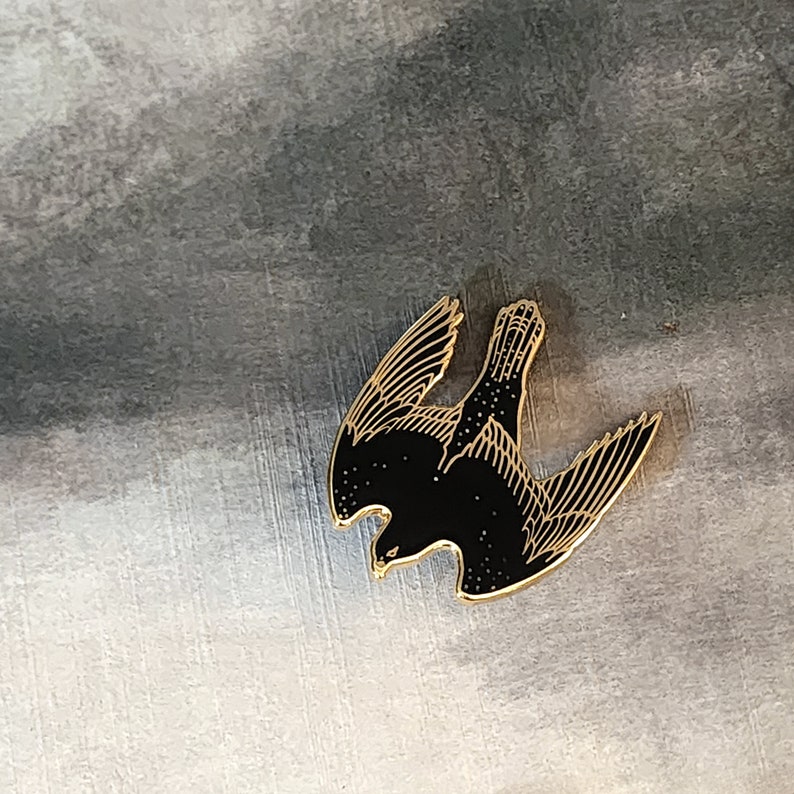 May include: A black and gold enamel pin featuring a stylized silhouette of a bird in flight.