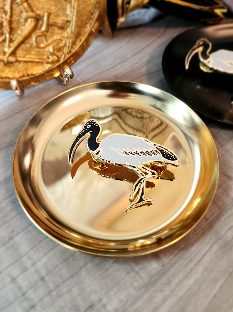 May include: A gold-colored metal tray with a white and black enamel pin of a bird with a long beak. The bird is standing on one leg and has its wings spread out.
