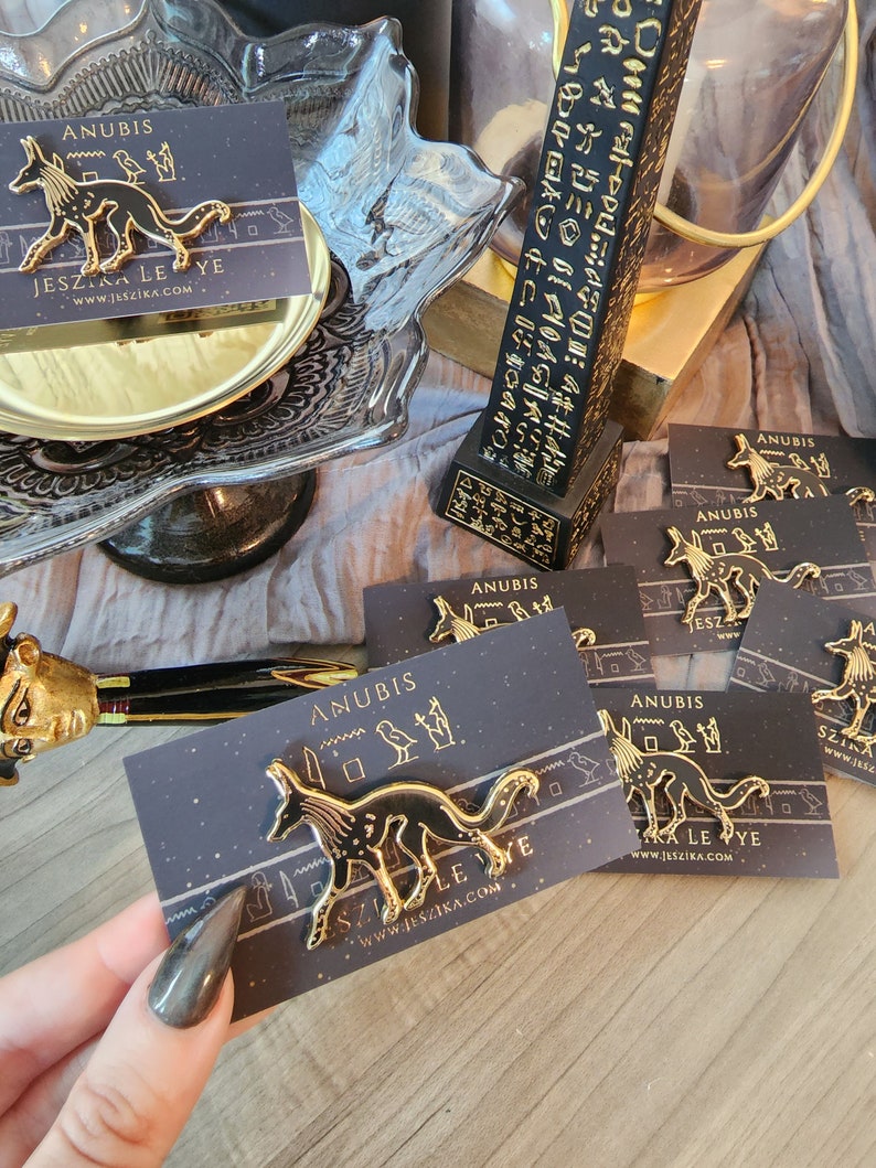 May include: A gold-colored enamel pin featuring a black and white stylized image of Anubis, the Egyptian god of the dead, with the text "Anubis" and "Jeszika Le Ye" and the website address "www.jeszika.com".