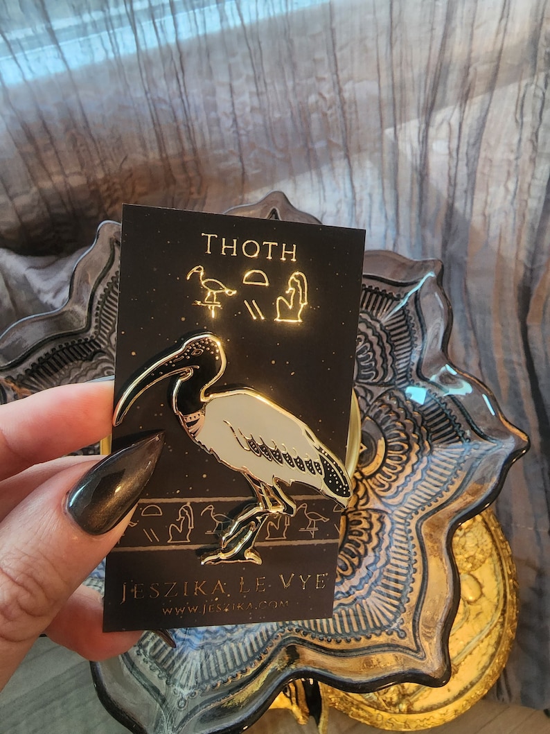 May include: Gold-toned enamel pin featuring a white ibis with black markings. The pin is on a black card with the text "THOTH" and "JESZIKA LE VYE" in gold lettering.