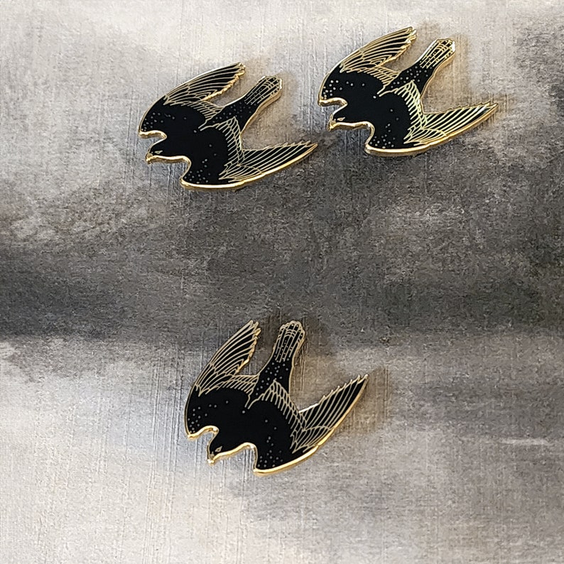 May include: Three gold-colored pins with black silhouettes of birds in flight. The birds are stylized and have a vintage aesthetic.