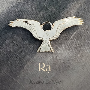 May include: A gold-colored metal pin of a white hawk with its wings spread wide. The pin has a loop at the top for attaching to clothing. The text "Ra" and "by Jeszika Le Vye" is below the pin.
