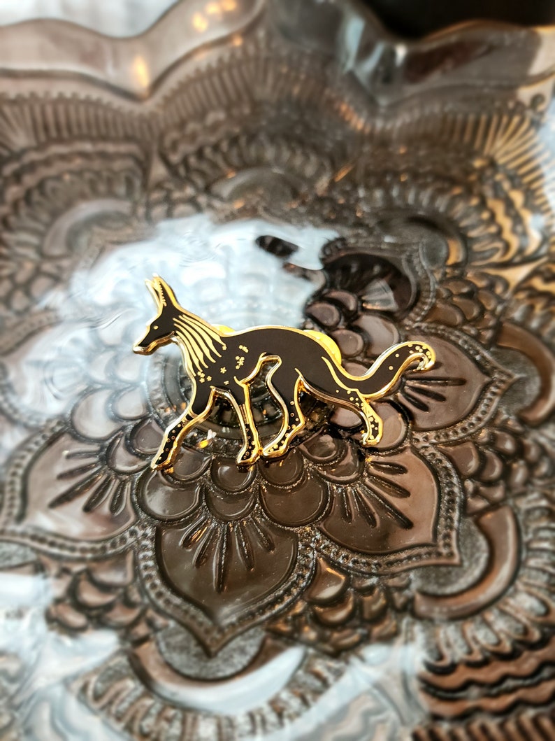 May include: Black and gold enamel pin featuring a stylized animal with a long, pointed snout and a long, flowing tail. The pin is set against a background of a decorative, mandala-like pattern.