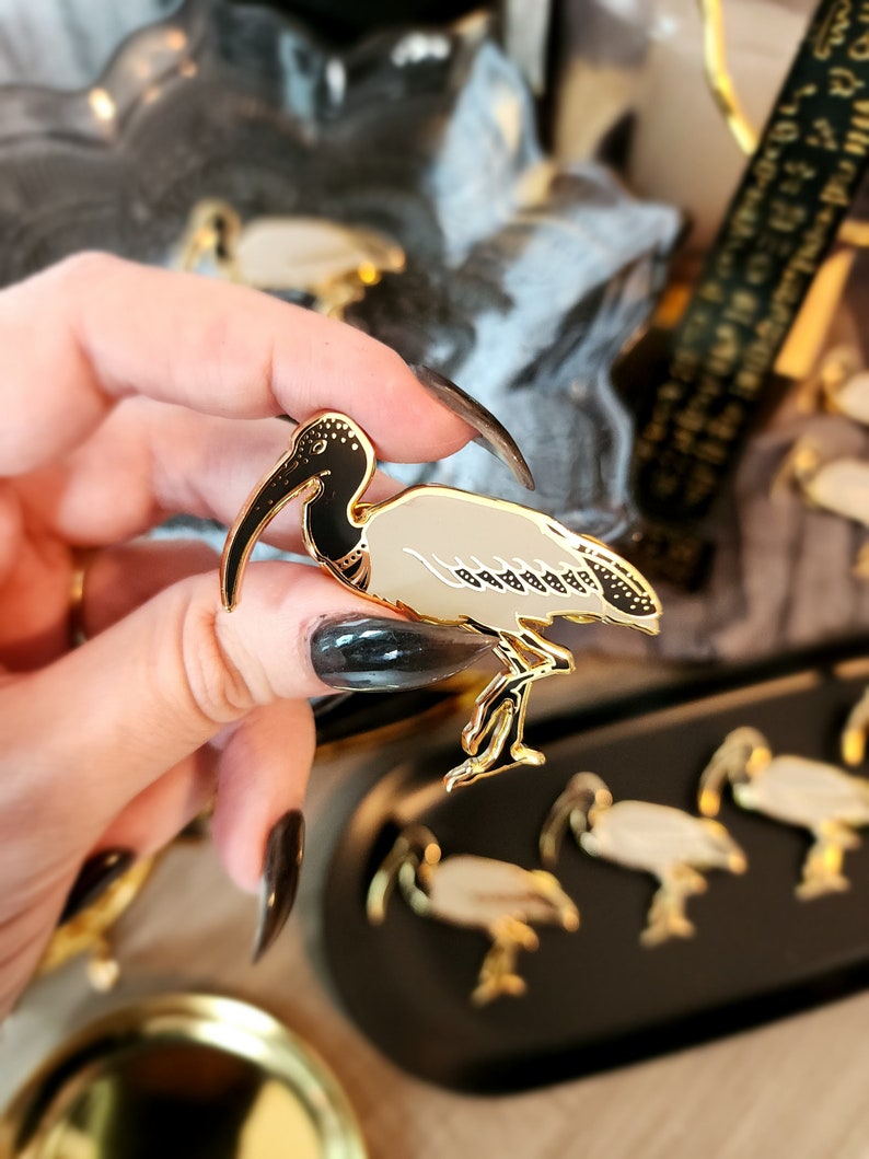 May include: A gold-tone enamel pin featuring a black and white ibis bird with a long, curved beak. The pin is being held by a hand with black nail polish.