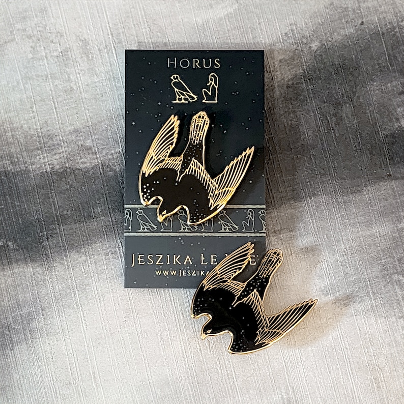 May include: Two gold-colored enamel pins featuring a black silhouette of a falcon with wings spread. The pins are on a black card with white text that reads "HORUS" and "JESZIKA LE" with a website address.