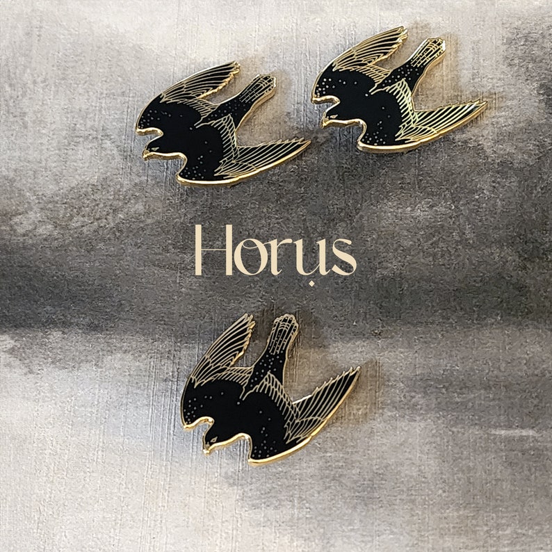 May include: Three gold-colored pins with black silhouettes of birds in flight. The word "Horus" is printed in gold below the pins.
