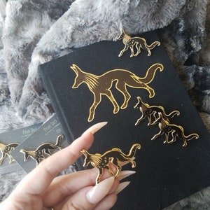May include: Five gold-colored enamel pins featuring a stylized fox design with black and gold details. The pins are arranged on a black surface with the text "From Dark to the" visible in the background.