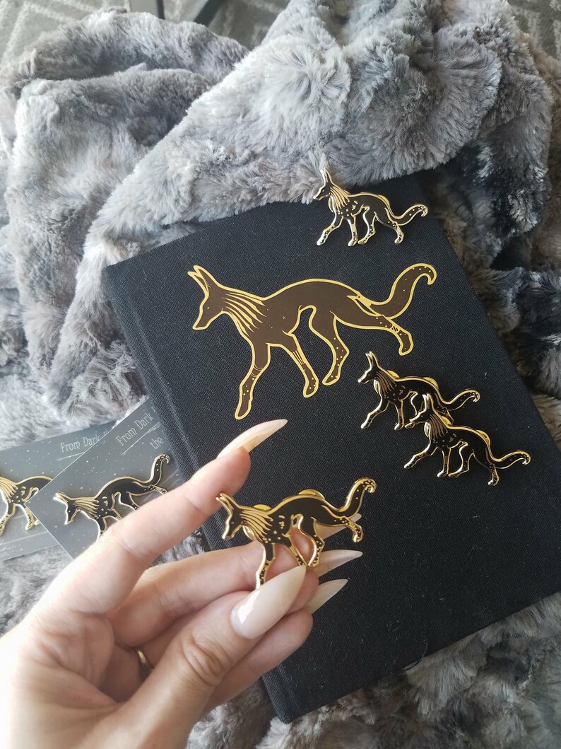 May include: Five gold-colored enamel pins featuring a stylized fox design with black and gold details. The pins are arranged on a black surface with the text "From Dark to the" visible in the background.