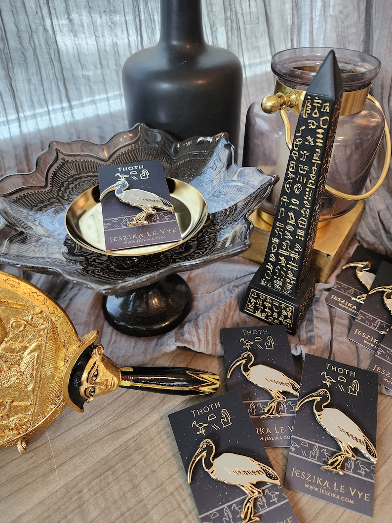 May include: Four gold-colored enamel pins featuring a white ibis bird with black markings. The pins are on black cards with gold text that reads "THOTH" and "JESZIKA LE VYE".