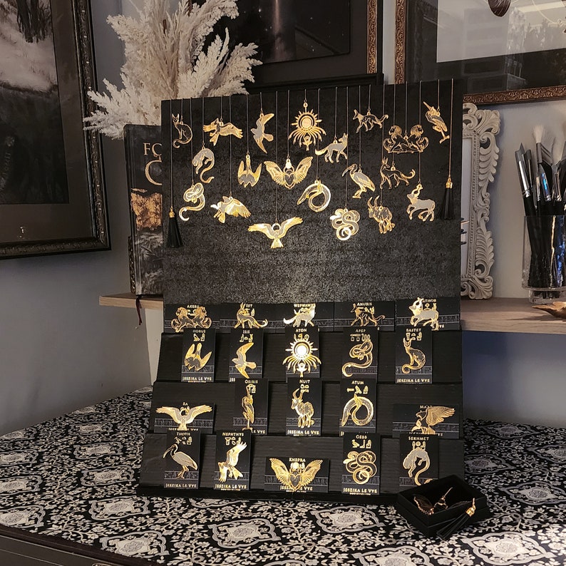 May include: A collection of 20 gold-toned metal charms featuring various animal and geometric designs. The charms are displayed on a black background with a wooden shelf below.
