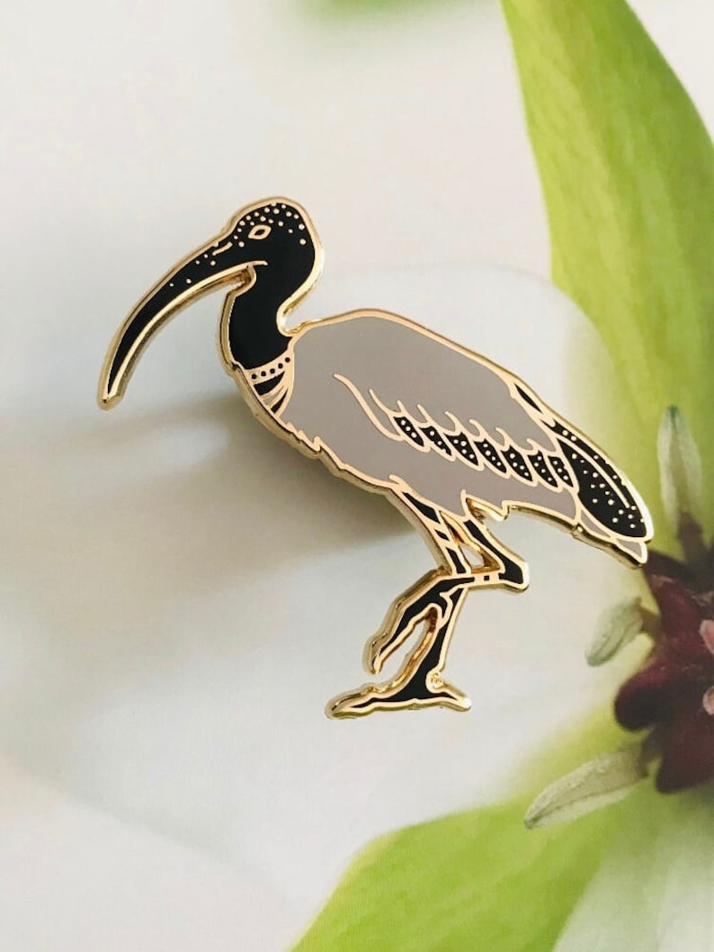 May include: A gold-tone enamel pin featuring a grey and black ibis bird with a long, thin beak. The bird is standing on one leg with its wings folded.