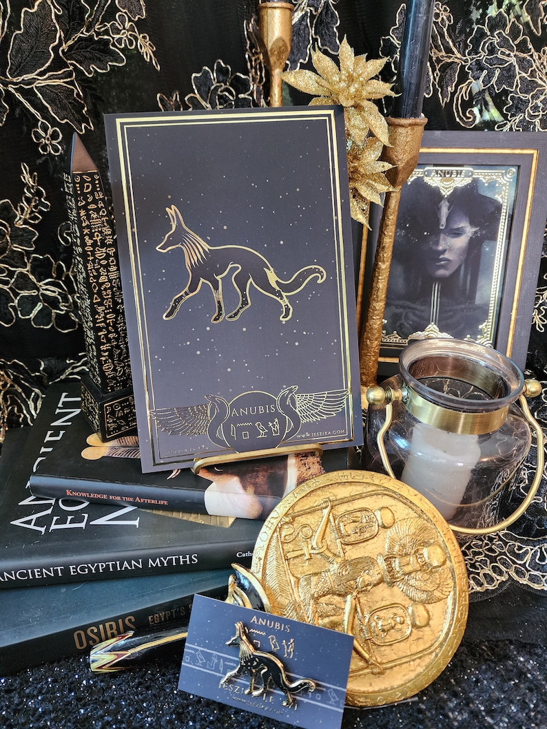 May include: A black and gold book with a stylized image of Anubis, the Egyptian god of the dead, on the cover. The book is surrounded by other Egyptian-themed items, including a gold coin with a relief of Anubis, a gold candle holder, and a book on ancient Egyptian myths.