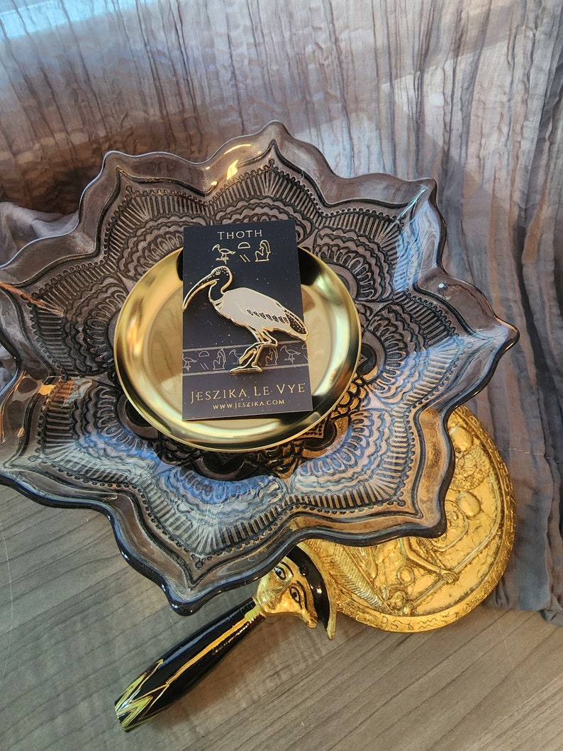May include: A gold pin featuring a white ibis with the text "THOTH" and hieroglyphs. The pin is on a gold plate with a black background. The text "JESZIKA LE VYE" and "WWW.JESZIKA.COM" are on a black card below the pin.