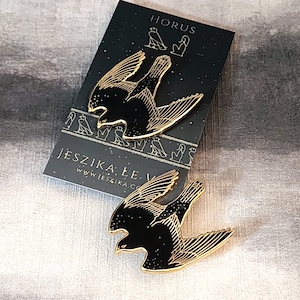 May include: Two gold-colored enamel pins depicting a black silhouette of a falcon with its wings spread. The pins are on a black background with gold text that reads "HORUS" and "JESZIKA LE" with a website address.