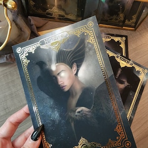 May include: A tarot card with a gold ornate frame and a black background. The card features a woman with a hawk on her arm and the word "HORUS" in gold lettering.