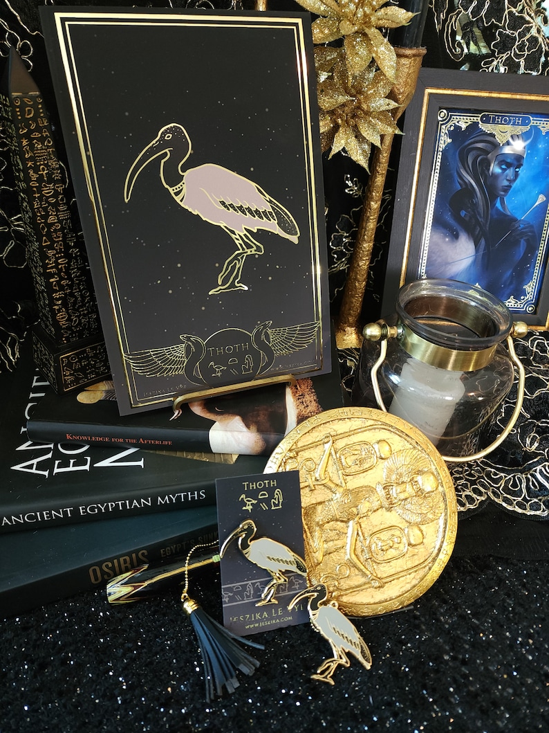 May include: Gold and black card with a gold ibis bird illustration and the word "THOTH" in hieroglyphs. The card is on a black surface with a gold book and a gold decorative plate with a hieroglyphic design.