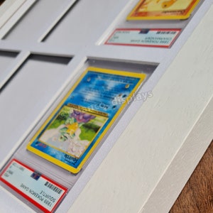 May include: White wooden frame with a white mat and glass panels. The frame holds multiple trading cards, including a Squirtle card from the 1999 Pokemon Game.