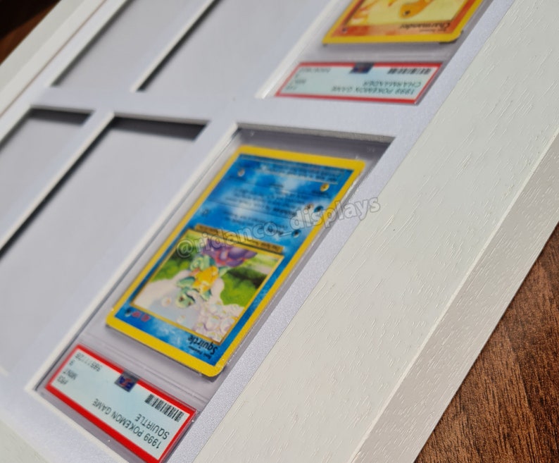 May include: White wooden frame with a white mat and glass panels. The frame holds multiple trading cards, including a Squirtle card from the 1999 Pokemon Game.