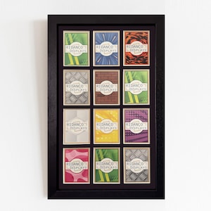 Op de afbeelding: Een zwart omkaderd display met twaalf kaarten, elk met een uniek ontwerp en de tekst "RIDANCO DISPLAYS". De kaarten tonen verschillende patronen en kleuren, gerangschikt in een raster binnen het frame.