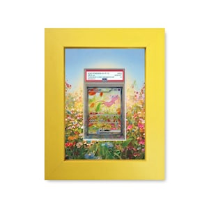 May include: A framed Pokemon card featuring a pink Mew character, set against a colorful floral background. The card is encased in a clear protective holder and displayed within a bright yellow frame. The frame is rectangular.