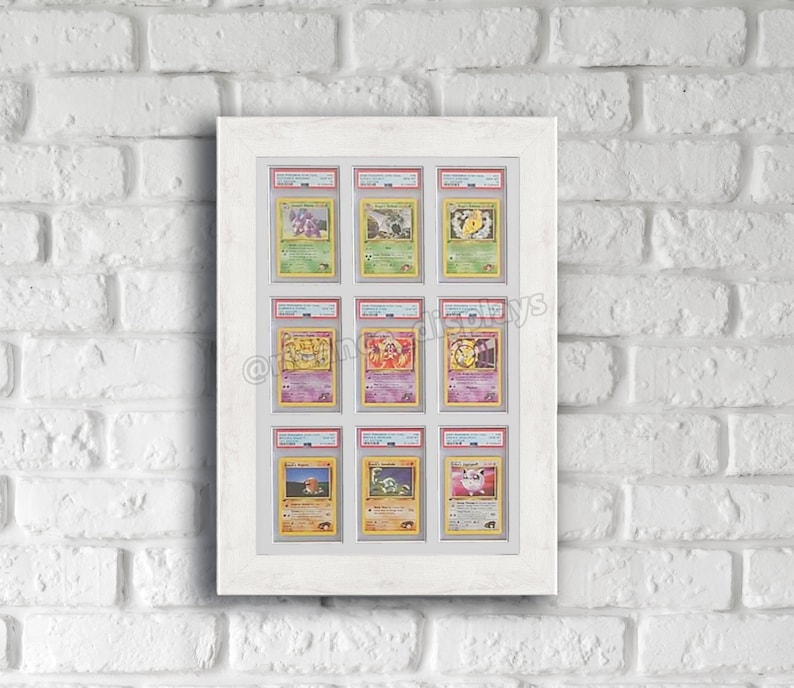 May include: A white frame with a white mount displays nine Pokemon cards in protective plastic sleeves. The cards are arranged in three rows of three. The cards are all from the first edition of the Pokemon Trading Card Game.