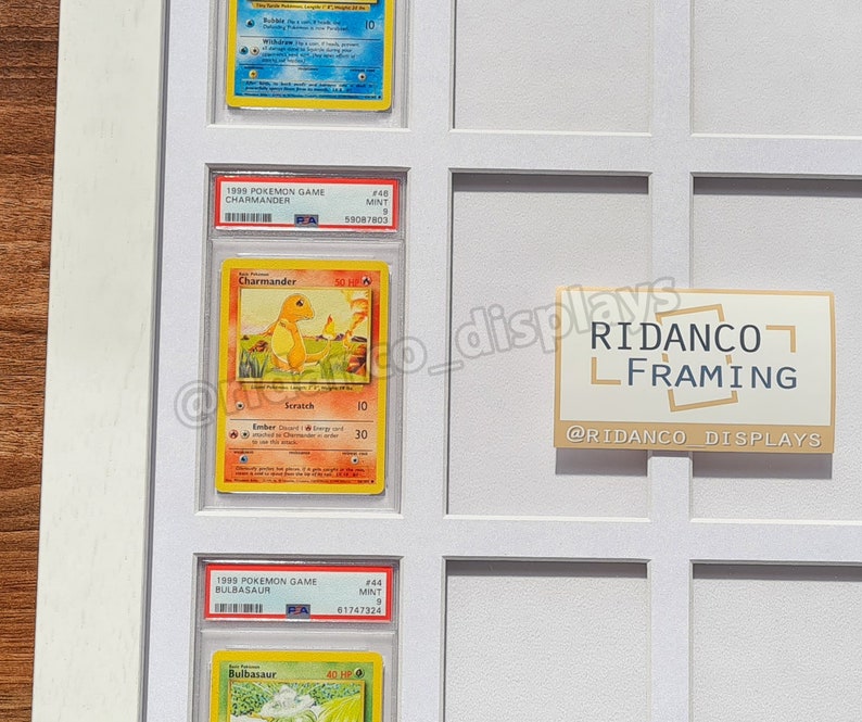 May include: A white display case with three Pokemon cards inside. The cards are from the 1995 and 1999 Pokemon Game series. The cards are Charmander, Bulbasaur, and a third card that is not visible. The display case has a white background and a black border. The text on the display case reads "RIDANCO FRAMING" and "@RIDANCO_DISPLAYS".