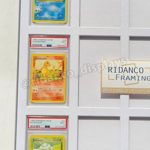 May include: A white display case with three Pokemon cards inside. The cards are from the 1995 and 1999 Pokemon Game series. The cards are Charmander, Bulbasaur, and a third card that is not visible. The case has a white background and a black border. The case is labeled "RIDANCO FRAMING" with the text "@RIDANCO_DISPLAYS" below.