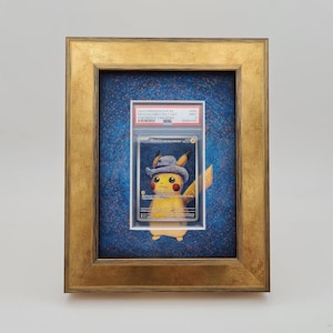 Op de afbeelding: Een ingelijste Pokemon-verzamelkaart met Pikachu die een blauwe cowboyhoed draagt. De kaart is beoordeeld en zit in een beschermende plastic hoes. De lijst is goud met een blauwe achtergrond.