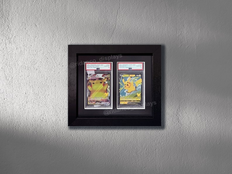 May include: Two Pokemon trading cards, a Pikachu and a Raichu, are displayed in a black frame with a white mat. The cards are in a protective plastic sleeve. The frame is on a gray wall. The text "@ridasco_displays" is visible in the bottom right corner of the image.