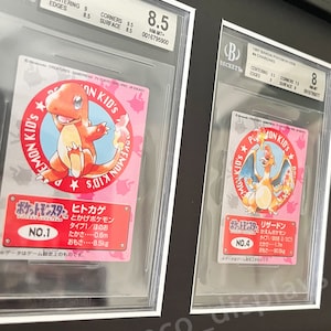 May include: Two Pokemon trading cards graded by Beckett Grading Services. The cards are both from the 1997 Bandai Pokemon Kids set. The first card is a Charmander, graded NM-MT+ 8.5. The second card is a Charizard, graded 8. Both cards feature the Pokemon in a circular frame with Japanese text.