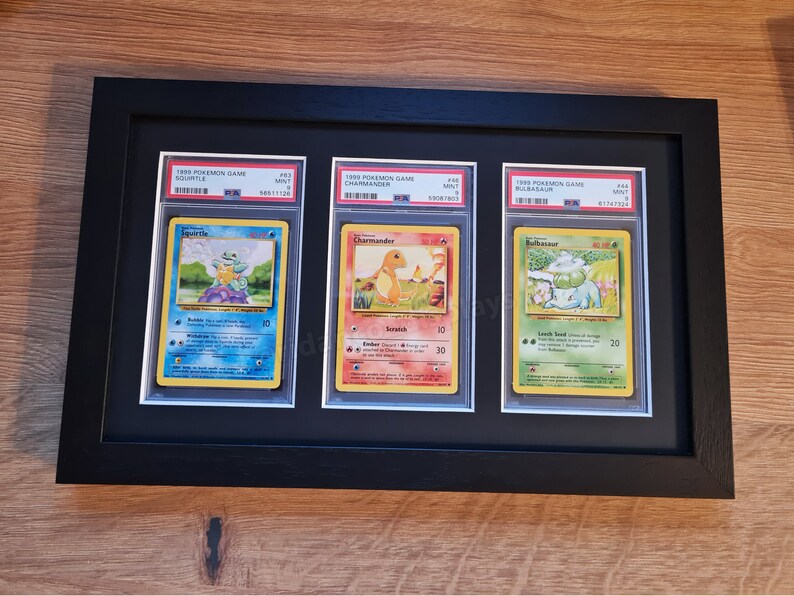May include: Three Pokemon trading cards, Squirtle, Charmander, and Bulbasaur, are displayed in a black frame. The cards are from the 1999 Pokemon Game series and are in mint condition.