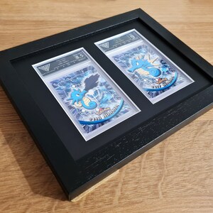May include: Two Pokemon trading cards, a Seadra and a Kingdra, are displayed in a black frame with a white mat. The cards are graded 8.5 and 5, respectively. The cards are in a protective plastic sleeve.