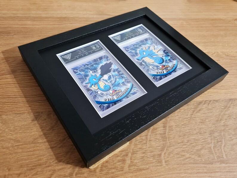 May include: Two Pokemon trading cards, a Seadra and a Kingdra, are displayed in a black frame with a white mat. The cards are graded 8.5 and 5, respectively. The cards are in a protective plastic sleeve.