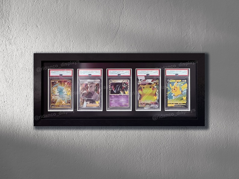 Op de afbeelding: Vijf Pok&eacute;mon-verzamelkaarten worden tentoongesteld in een zwart frame met een witte passe-partout. De kaarten zijn ingekapseld in beschermende plastic hoesjes. De kaarten tonen Pikachu, Charizard en andere Pok&eacute;mon-personages. Het frame is gelabeld met de social media-handle "@ridanco_displays".