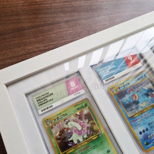 May include: A white frame with a glass front displays four Pokemon cards. The cards are graded and labeled with their condition and number. The cards are all from the 1999 Pokemon Trading Card Game.