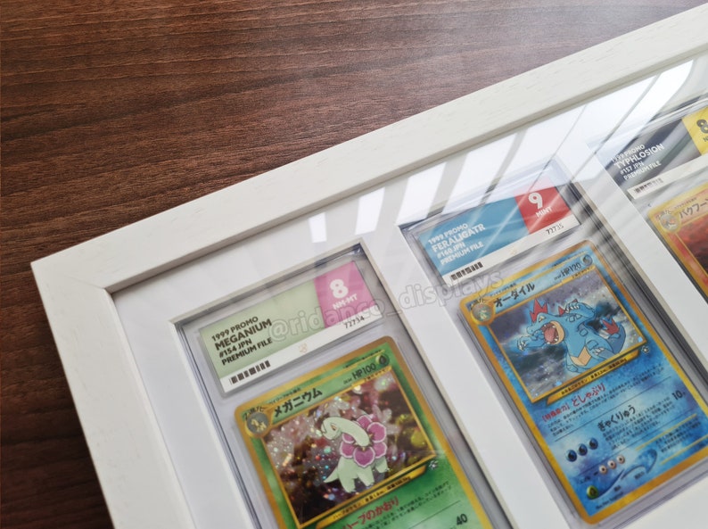 May include: A white frame with a glass front displays four Pokemon cards. The cards are graded and labeled with their condition and number. The cards are all from the 1999 Pokemon Trading Card Game.