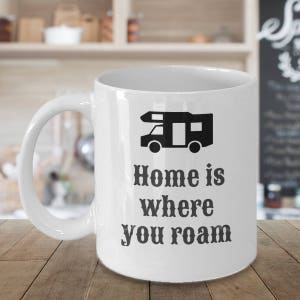 以下が含まれることがあります： 白いセラミックマグカップ。黒いキャンピングカーのグラフィックと「Home is where you roam」のテキストが特徴です。 マグカップは湾曲したハンドルを持ち、木製の表面に置かれています。