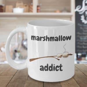 以下が含まれることがあります： 白いセラミック製のマグカップに、茶色の棒人間が白いマシュマロを3つ持っています。マグカップには「marshmallow addict」という文字が印刷されています。