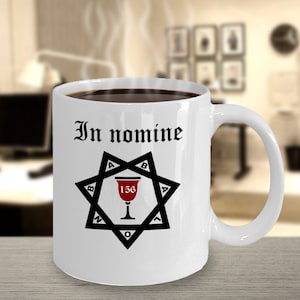 May include: White ceramic coffee mug with a black and red design. The mug features a six-pointed star with a red chalice in the center. The text "In nomine" is printed above the star.