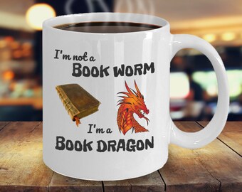 Op de afbeelding: Een witte keramische koffiemok met een zwarte handgreep. De mok heeft een zwarte en oranje afbeelding van een draak en een boek met de tekst "I'm not a book worm I'm a book dragon".