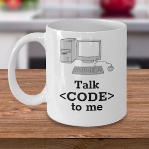 Op de afbeelding: Witte keramische mok met een zwarte omtrek van een computer, toetsenbord en muis. De tekst "Talk <CODE> to me" staat op de mok gedrukt.