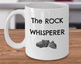 May include: White ceramic coffee mug with a black inscription that reads "The ROCK WHISPERER" and a small image of three gray rocks.