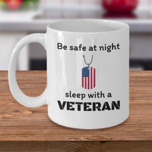 May include: White ceramic coffee mug with a black and white graphic that reads "Be safe at night sleep with a VETERAN". A silver chain with a dog tag with an American flag is hanging from the text.