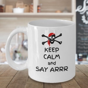 May include: White ceramic coffee mug with a black and red pirate skull and crossbones design. The mug has the text "Keep Calm and Say Arrr" printed on it.