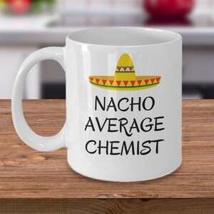 May include: White ceramic mug with a sombrero graphic and the text "NACHO AVERAGE CHEMIST" in black. The sombrero is yellow with red and green accents. The mug has a curved handle and is on a wooden surface.