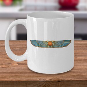 May include: White ceramic coffee mug featuring a blue and gold winged sun symbol. The symbol is a stylised representation of the ancient Egyptian sun god Ra.