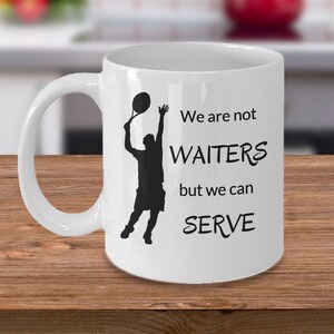 May include: White ceramic coffee mug with a black silhouette of a tennis player serving a ball. The mug has the text "We are not waiters but we can serve" printed on it.